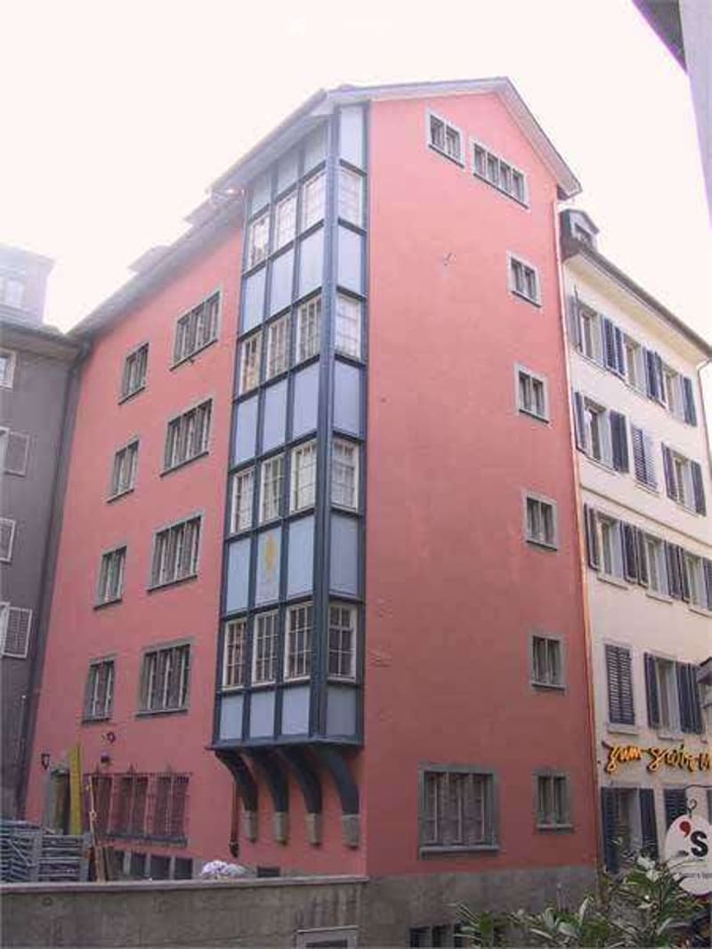 Multi-story building with red brick facade and numerous windows, featuring a prominent bay window with decorative wooden framing.