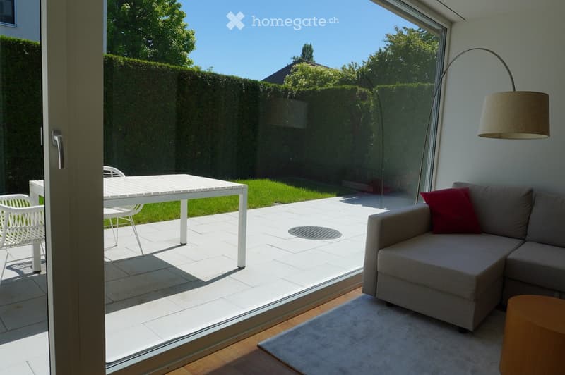 Modern living room with large glass doors opening to a patio, featuring a grey sectional sofa, a small wooden stool, a white outdoor table, and white wicker chairs.