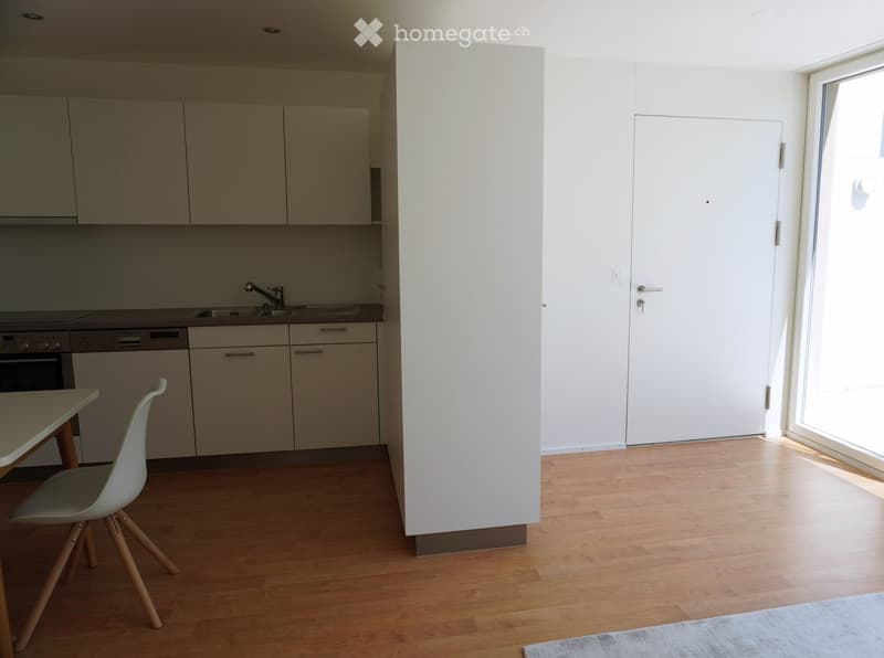 Modern kitchen with light-colored cabinets, wooden flooring, a small table with a single chair, and built-in appliances including an oven and dishwasher.
