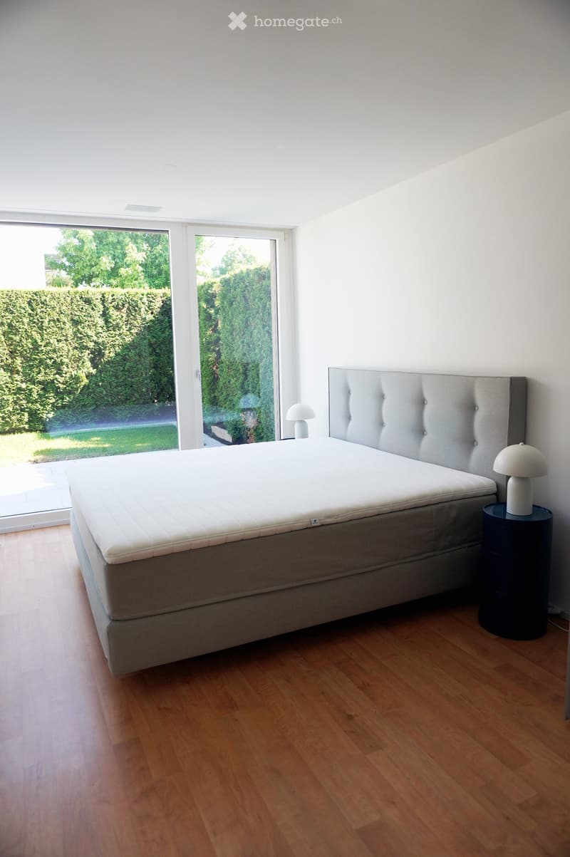 Modern bedroom with a large bed, tufted headboard, nightstand, and lamp. Large windows offer a view of greenery outside.