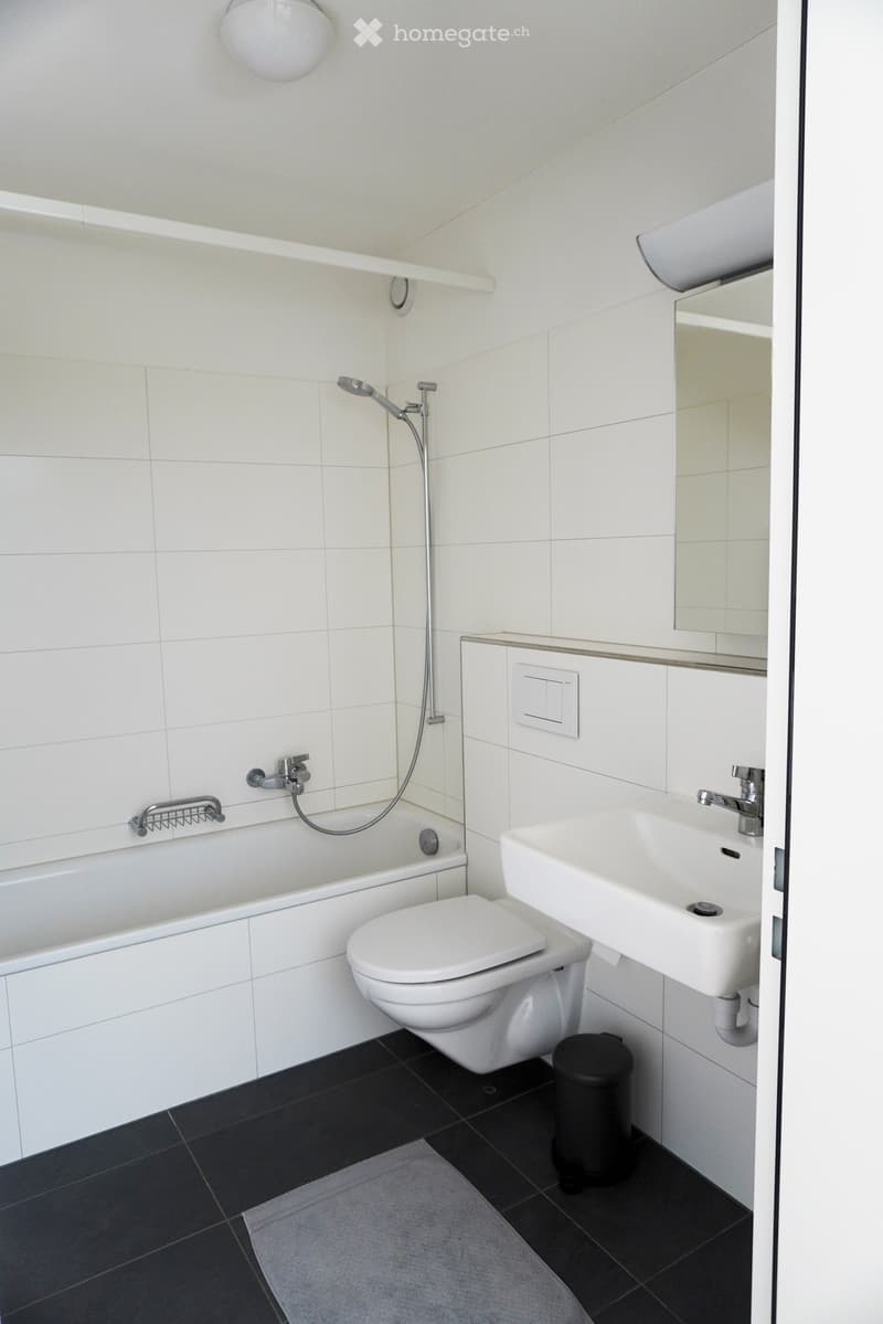 Modern bathroom with white tiles, bathtub, toilet, sink, mirror, and a light fixture above the sink.