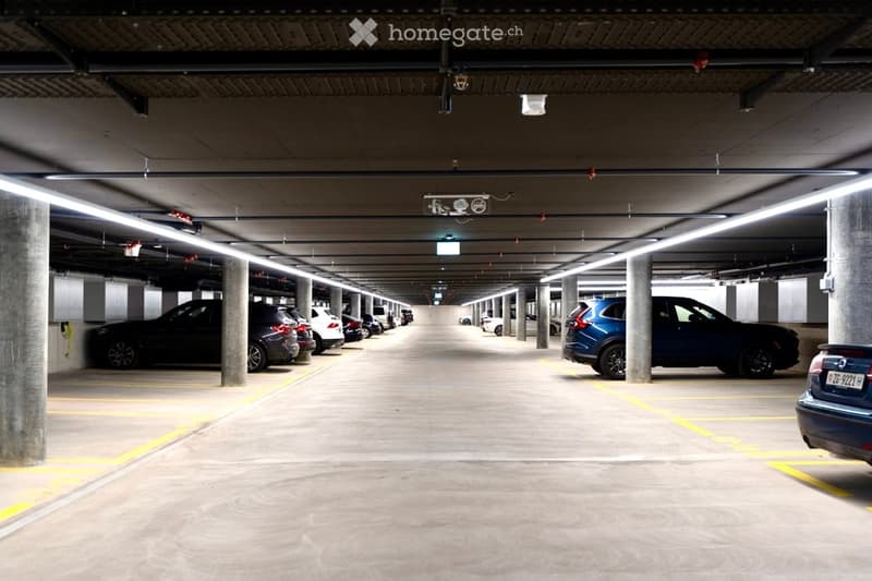 Underground parking area with multiple parked cars, concrete pillars, and overhead lighting.