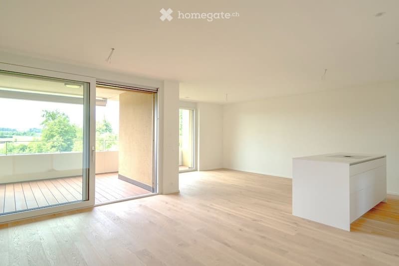 Bright, unfurnished living space with wooden flooring, large sliding glass doors leading to a balcony, and a white kitchen island.