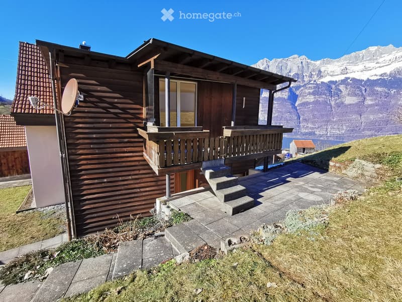 Two-story wooden house with a balcony, satellite dish, and steps leading to the entrance, set against a backdrop of mountains and a lake.