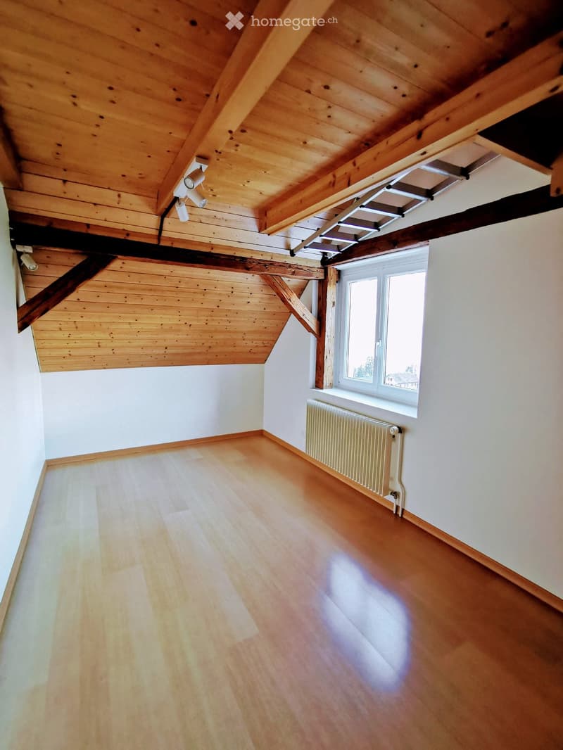 Empty attic space with wooden beams, exposed wooden ceiling, wooden floor, white walls, and a window with a radiator below it.