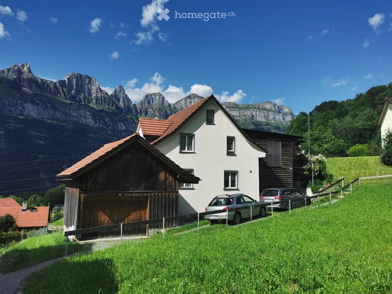 A two-story house with a red-tiled roof and white walls, featuring a wooden garage and a driveway with parked cars, set against a backdrop of mountains and greenery.