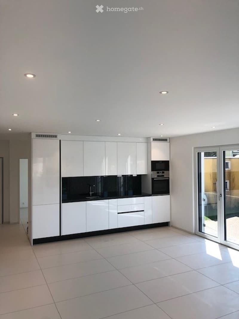 Modern, minimalist kitchen with white cabinets, black countertops, and built-in appliances including a sink, oven, and microwave. The space features recessed lighting and large tiles on the floor.