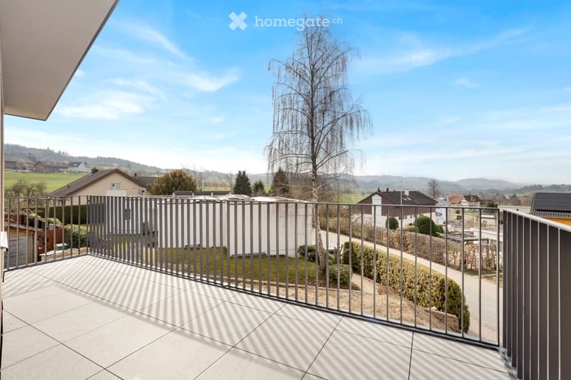 Balcony with tiled floor, metal railing, overlooking a residential area with houses, trees, and a clear blue sky.
