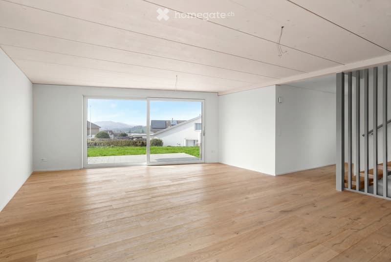 Empty room with light wooden flooring, white walls, and large glass doors leading to a view of a green lawn and distant houses.