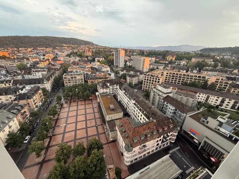 Aerial view of a cityscape with multiple buildings, streets, and a central square. The area includes a mix of residential and commercial structures, with visible rooftops and a distant hill.