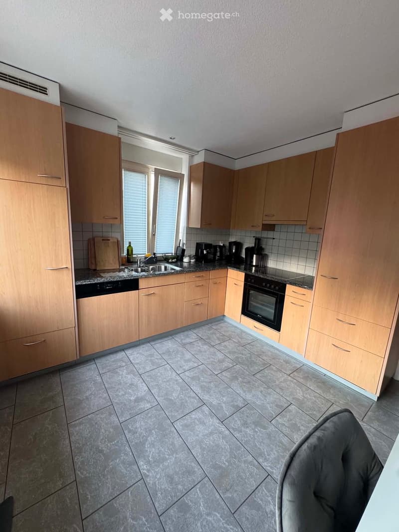 Modern kitchen with light wooden cabinets, granite countertops, and stainless steel appliances including a sink, oven, and dishwasher. The floor is tiled, and there is a window with blinds above the sink.