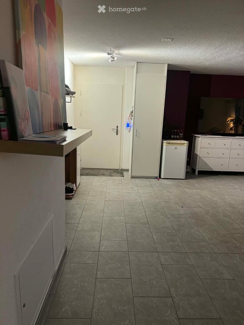 Empty hallway with tiled floor, white walls, and a closed door leading to another room. There is a small table with a few items on the left side and a white cabinet on the right side.