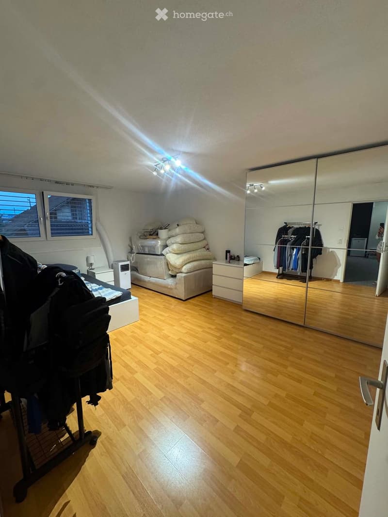 Bright bedroom with wooden flooring, large mirrored wardrobe, neatly made bed, and a stack of folded blankets on a bench.