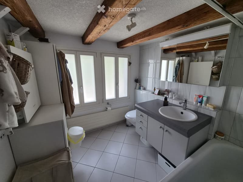 Bathroom with white tiled floor, wooden ceiling beams, sink with faucet, mirror, cabinet, toilet, and towel hanging on the wall.