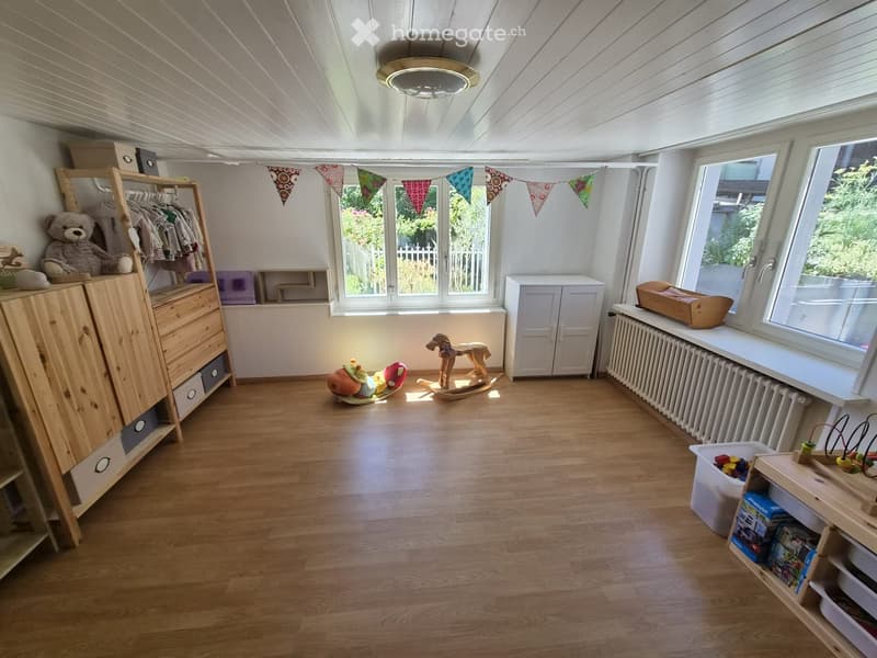 Brightly lit room with wooden flooring, large windows, and various children's furniture and toys including a wardrobe, toy horse, and shelves.