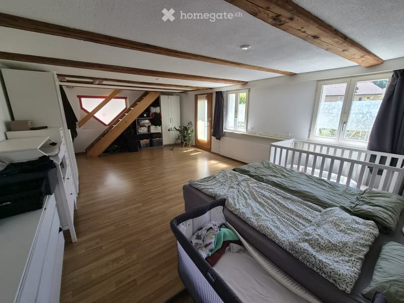 Bright, airy bedroom with wooden floors, exposed wooden beams, large windows, a sofa, a baby crib, a baby cot, a wardrobe, and a small plant near the window.