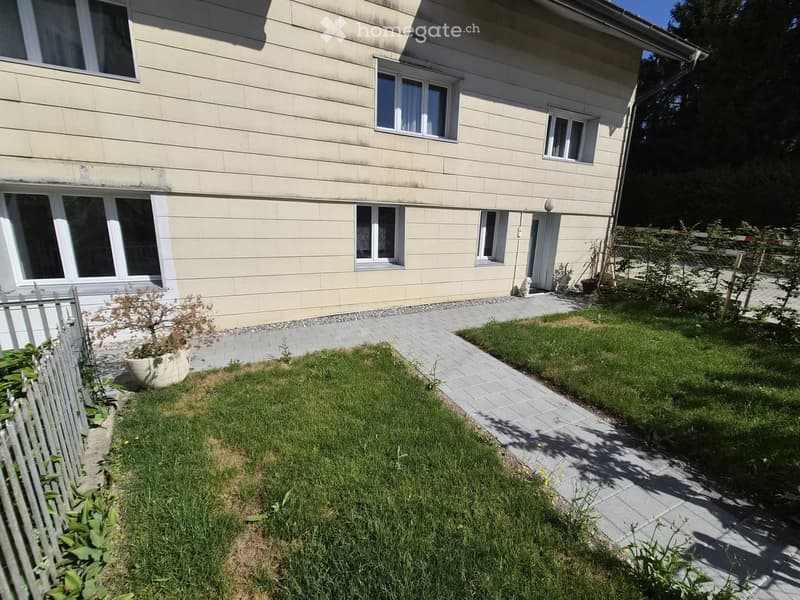 A well-maintained garden area with a paved pathway leading to a house entrance, featuring a small lawn, potted plants, and a white fence.