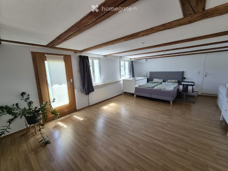 Bright bedroom with wooden flooring, exposed wooden beams, large windows, and a grey bed with matching nightstand. A small baby crib is visible in the background.