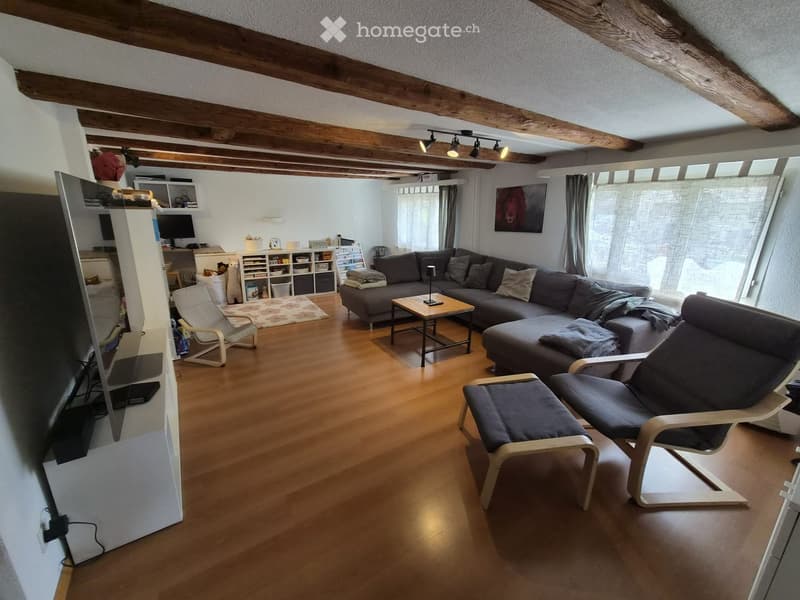 Living room with wooden floor, exposed wooden beams, large sectional sofa, coffee table, chairs, TV screen, and shelves.