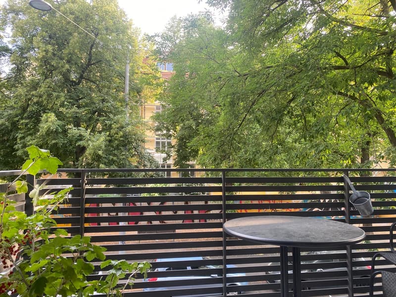Balcony with a round metal table and a chair, surrounded by greenery and a metal railing, overlooking a street with parked cars and trees.