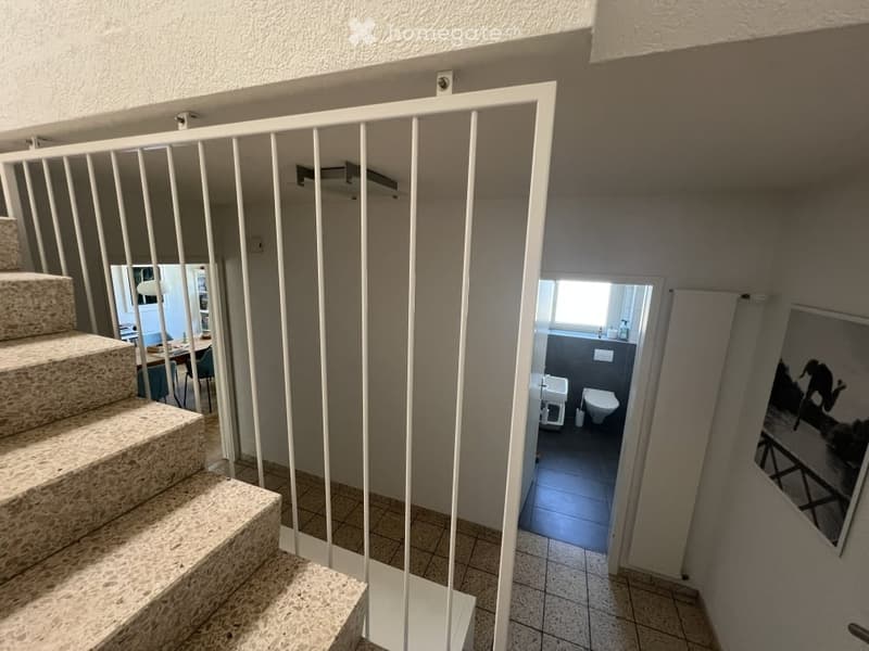 A modern hallway with a staircase leading upwards, white railing, and a view into a bathroom and dining area.