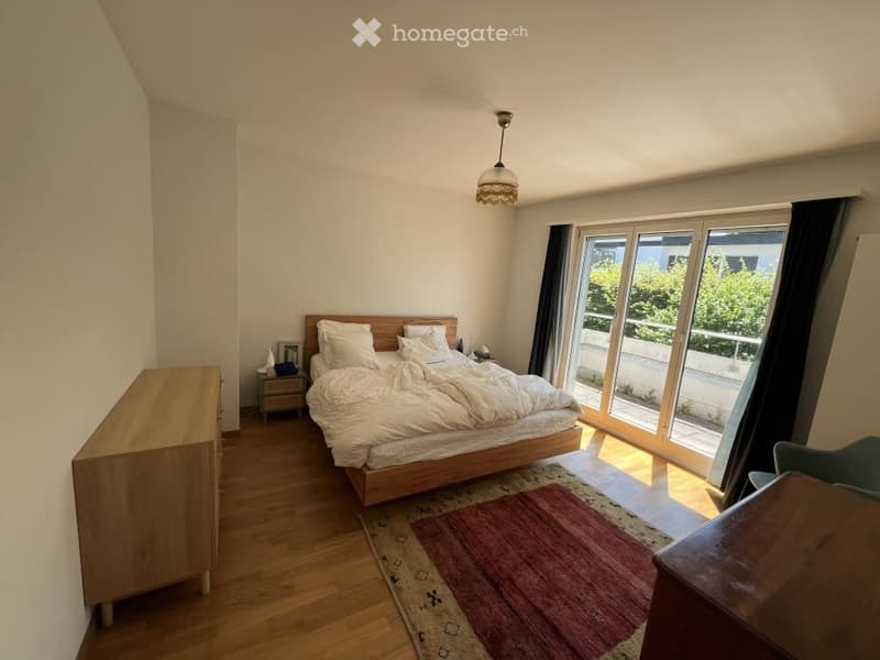 Bright bedroom with wooden flooring, large window with balcony access, bed with white bedding, nightstand, dresser, and a colorful rug.