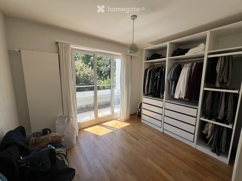 Bright room with wooden flooring, large window with balcony access, white wardrobe with multiple drawers and hanging space, and a hanging lamp.