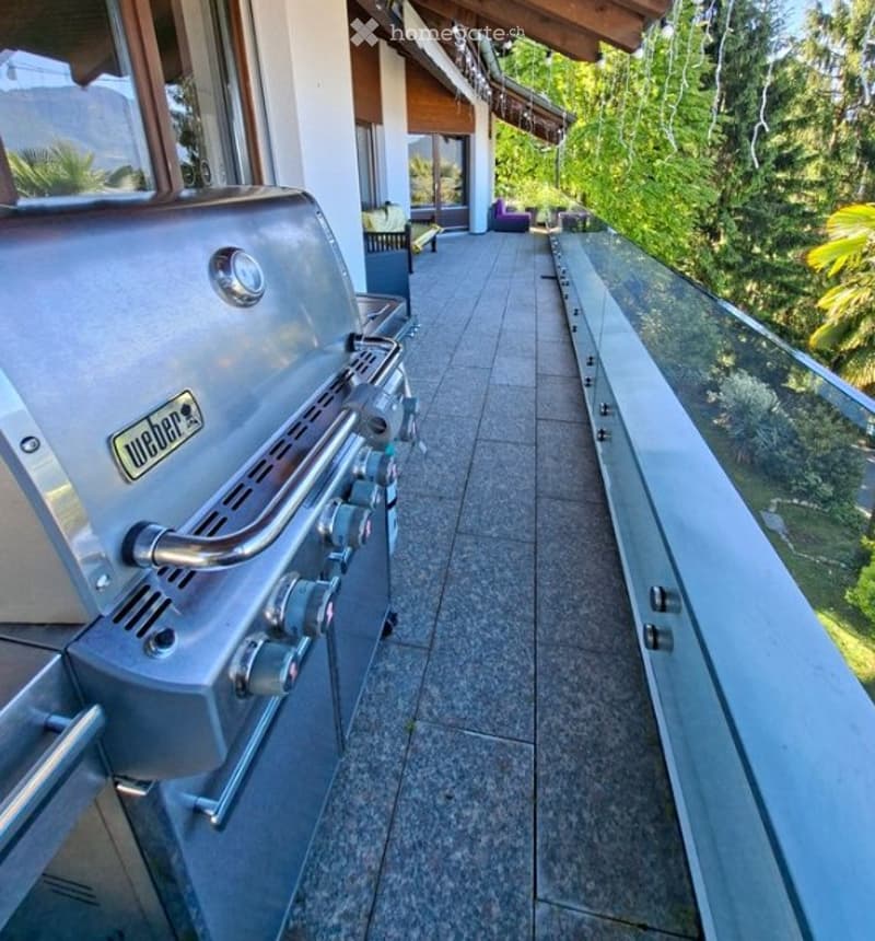 Modern balcony with a glass railing, tiled floor, and a Weber grill on the left side. The background shows a lush green view of trees and part of a house with outdoor seating.