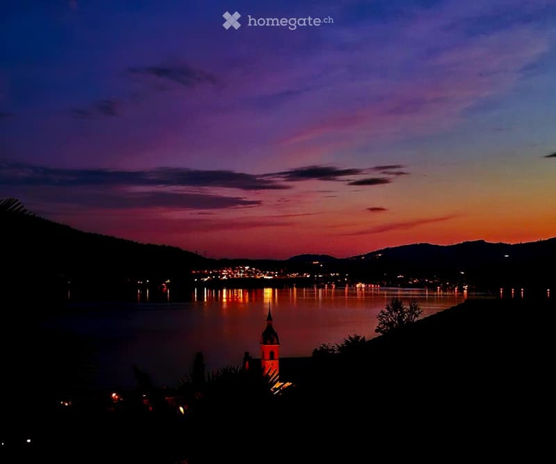 Evening view of a lake with city lights reflecting on the water, surrounded by hills and a small illuminated tower.