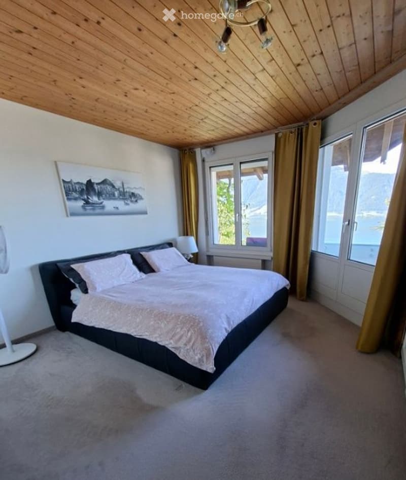 Modern bedroom with a large bed, nightstand, lamp, fan, and a scenic view through large windows with curtains.