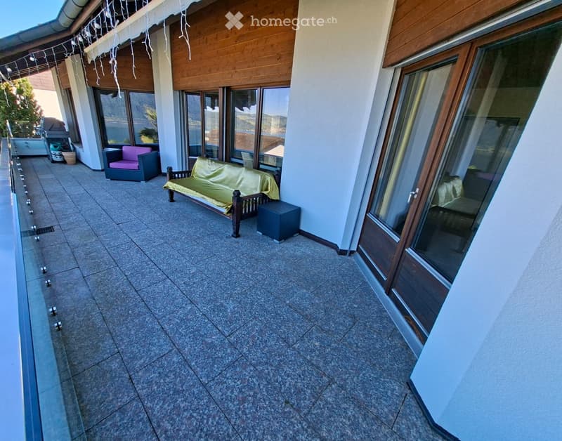 Balcony with tiled floor, large windows, sofa, chair, and bench, overlooking a scenic view.
