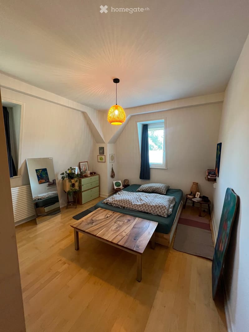 Cozy attic bedroom with wooden flooring, slanted ceiling, and large window. Features a bed with patterned bedding, a wooden coffee table, a mirror, a small cabinet with a plant, and wall decorations.