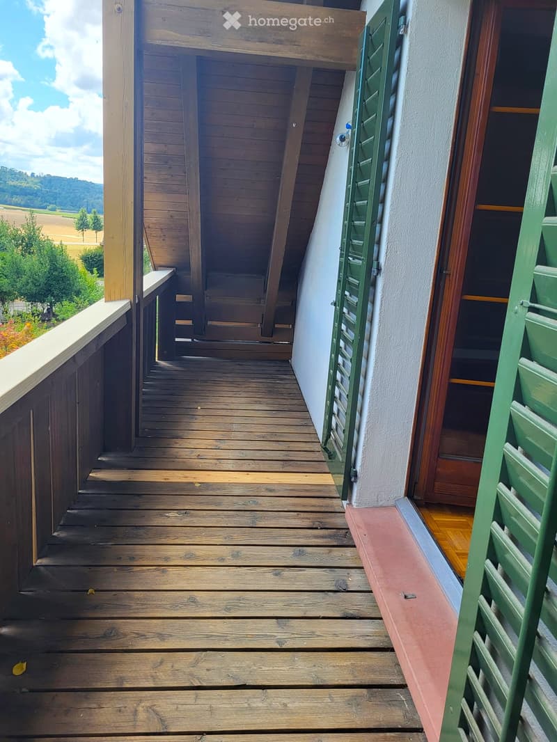 Wooden balcony with green shutters, overlooking a rural landscape with trees and fields.
