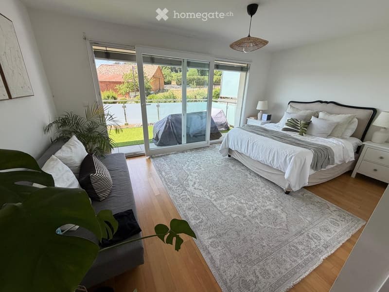 Bright bedroom with large windows and sliding glass doors leading to a balcony. Features a grey sofa, a large bed with white and grey bedding, a nightstand with a lamp, and a patterned rug on the wooden floor.