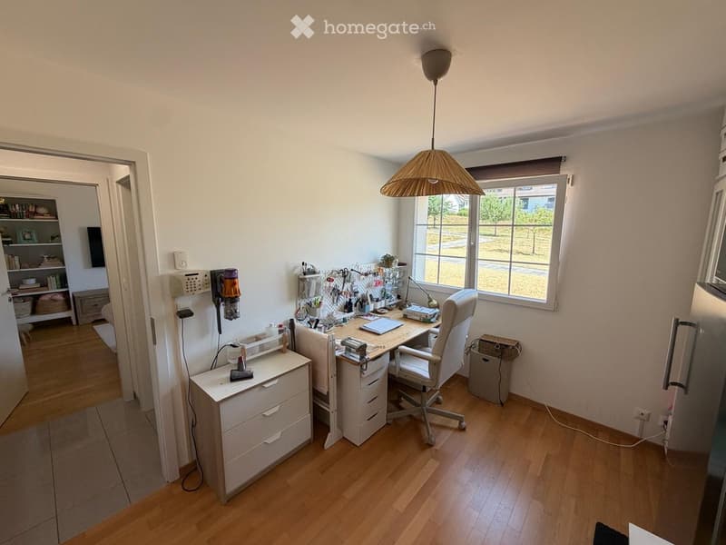 A well-lit home office with a wooden floor, a large window, a desk with a chair, a hanging lamp, a shelf with books and tools, and a refrigerator in the background.