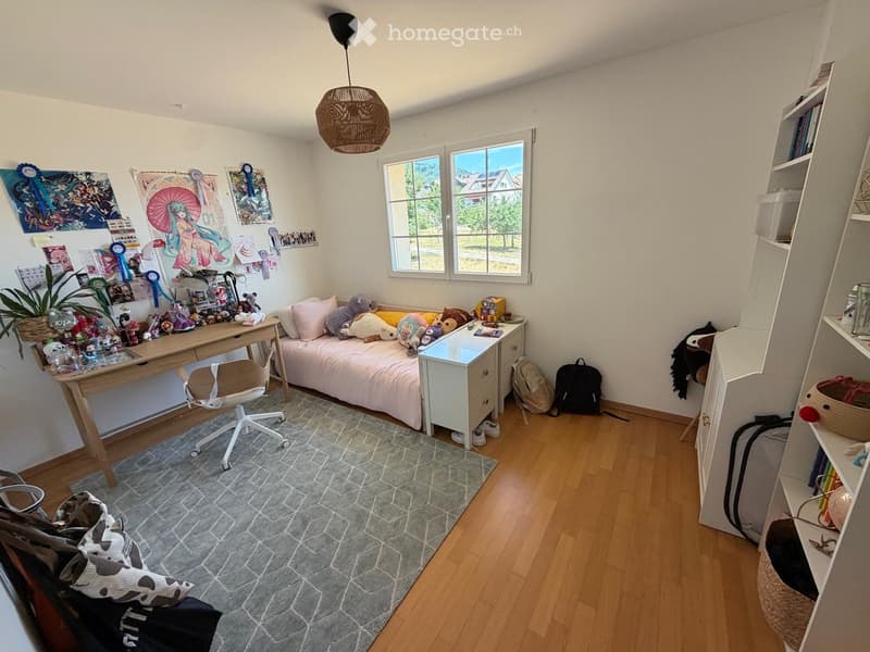 Cozy bedroom with a small bed, desk, chair, and shelves. Features a large window with a view, a woven hanging lamp, and various decorative items including posters and toys.