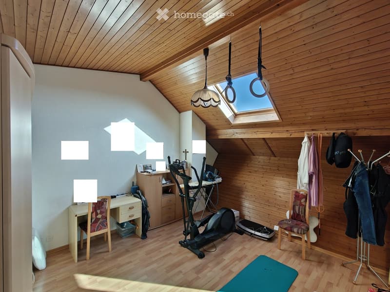 A well-lit room with wooden walls and ceiling, featuring a desk with a chair, a cabinet, exercise bike, treadmill, and a coat rack with hanging clothes.