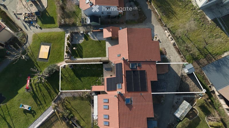Aerial view of a two-story house with red-tiled roof, solar panels on the roof, and a well-maintained lawn with a wooden deck and outdoor seating area.