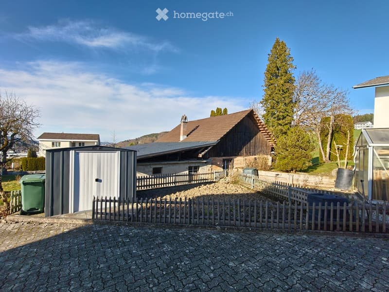 A garden area with a wooden fence, a shed, and a house with a brown roof in the background. The ground is paved with stone tiles, and there are trees and bushes around the area.