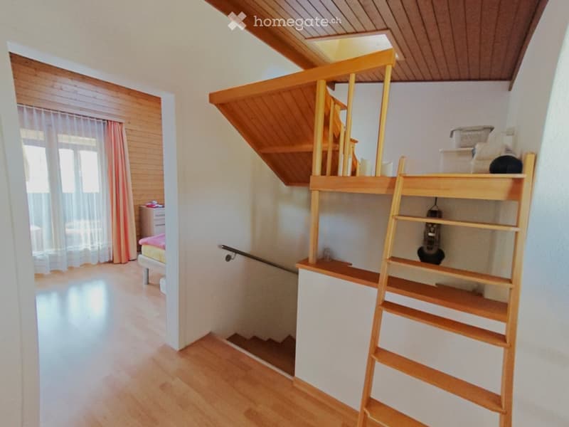 Cozy attic bedroom with wooden floors, slanted wooden ceiling, built-in loft bed, small window with curtains, and a wooden shelf unit.
