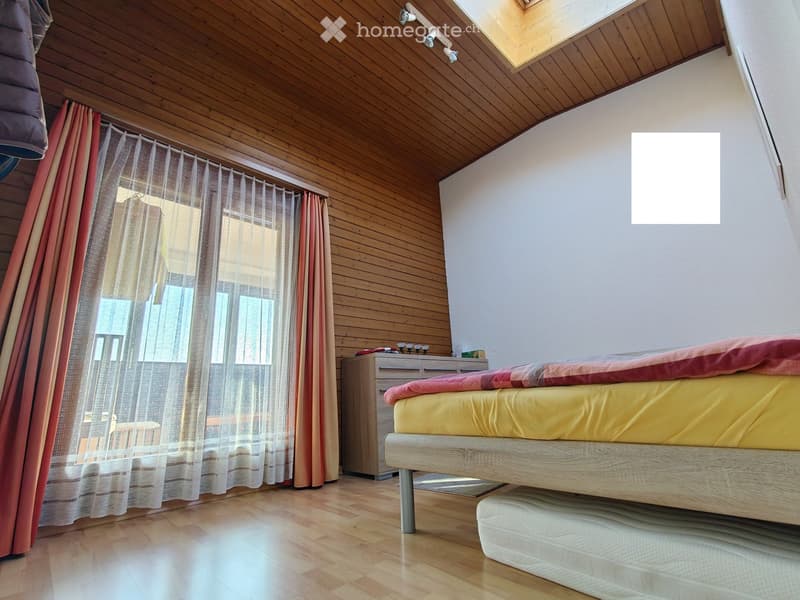 Bright bedroom with wooden ceiling and floor, large window with sheer curtains, bed with yellow and red bedding, nightstand with small items.