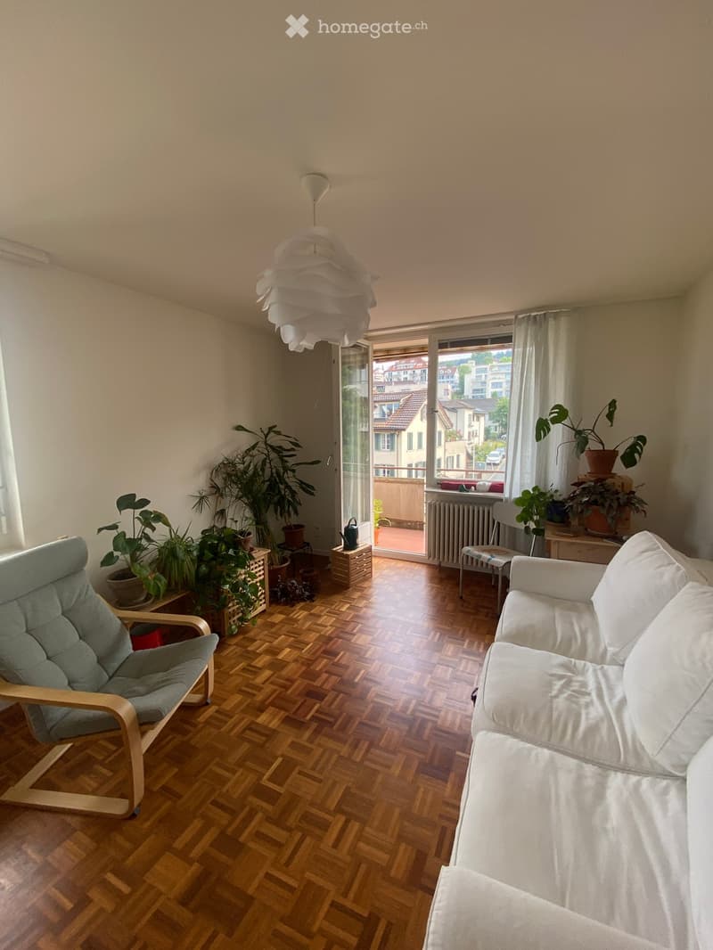 Living room with wooden flooring, white sofa, armchair, potted plants, and a view of the city through large windows.