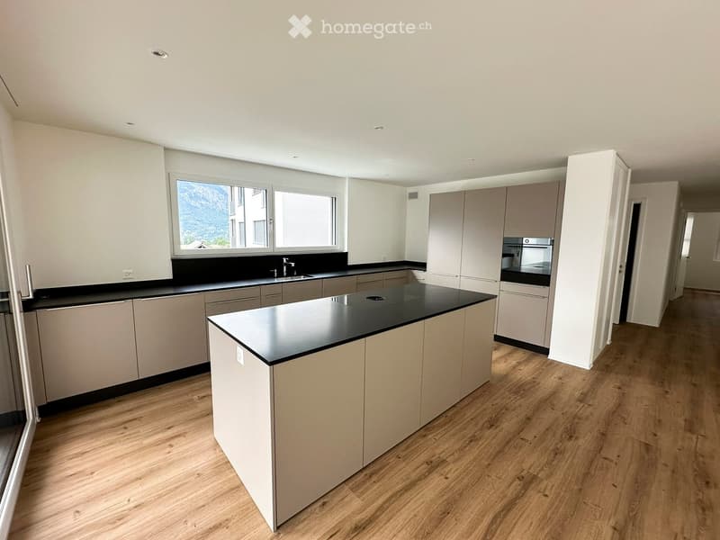 Modern kitchen with light-colored cabinets, black countertops, and wooden flooring. Features an island with a sink and an oven.