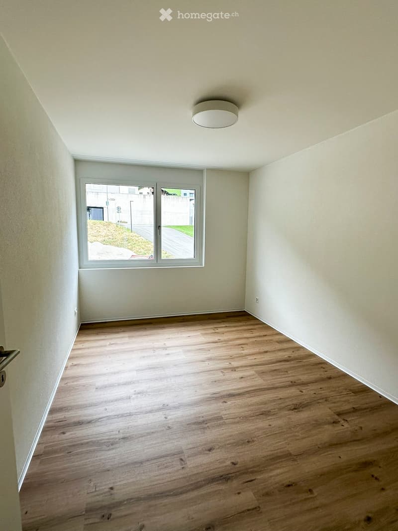 Empty room with light wooden flooring, white walls, and a large window with a view of the outside. A single ceiling light fixture is visible.