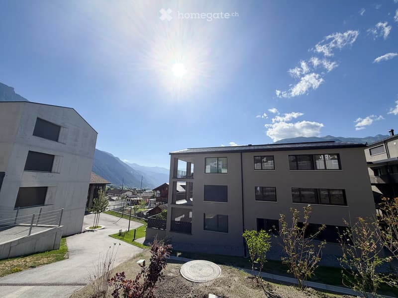 Modern two-story building with large windows and balconies, surrounded by greenery and a scenic mountain backdrop.