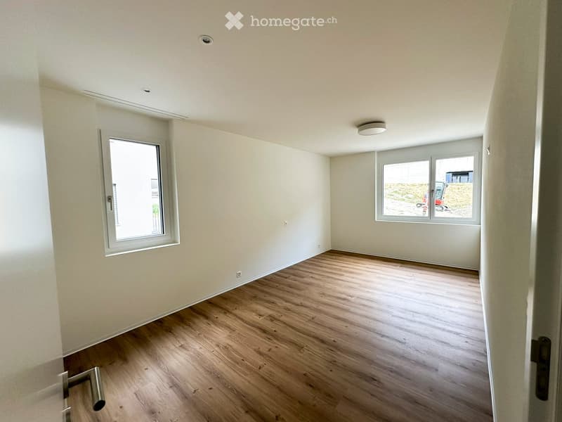 Empty room with light-colored walls, wooden flooring, and two windows allowing natural light. The room features a modern ceiling light fixture and a door frame visible on the right side.