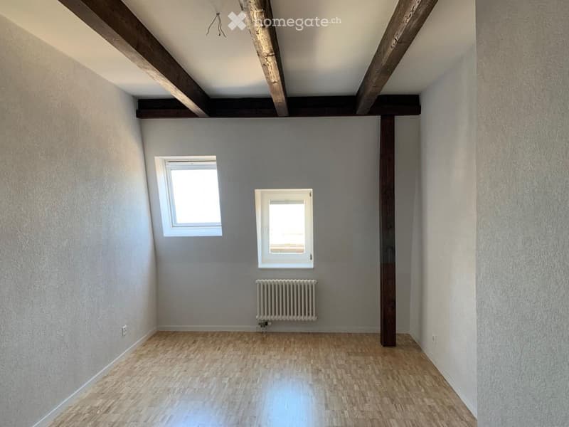 Empty room with wooden flooring, exposed wooden beams on the ceiling, two windows, and a radiator.