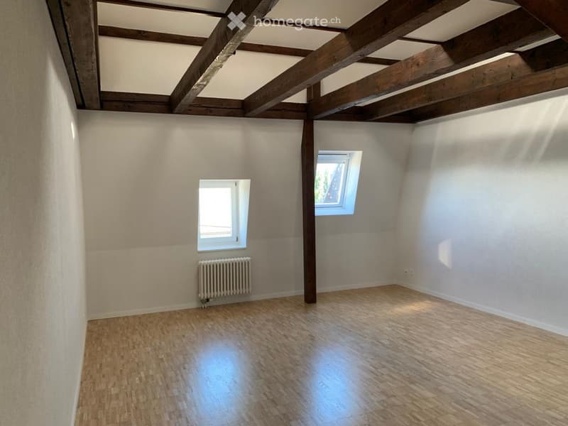 Empty room with wooden flooring, white walls, exposed wooden beams on the ceiling, and two small windows allowing natural light.