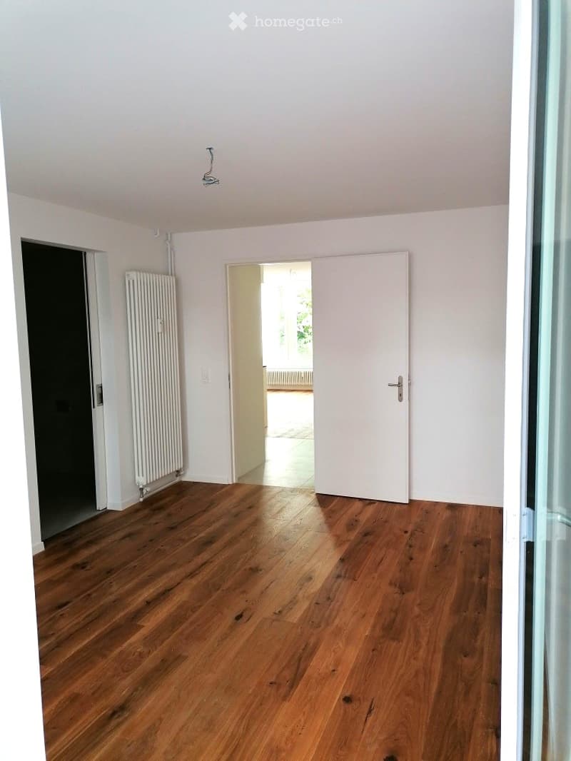 Empty room with wooden flooring, white walls, and a radiator on one wall. There are two doors, one leading to another room and the other to an outdoor area.
