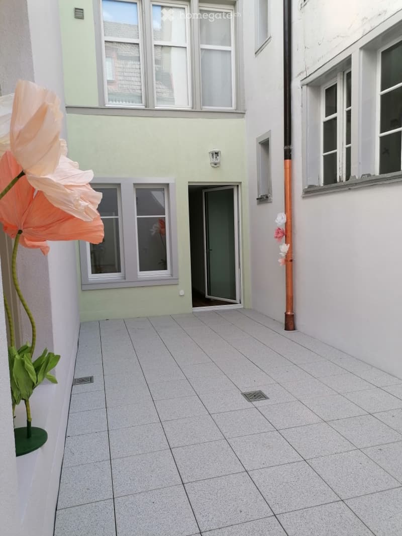 A small, enclosed patio area with light grey tiled flooring, surrounded by white walls and windows. A large pink flower in a pot is visible on the left side.